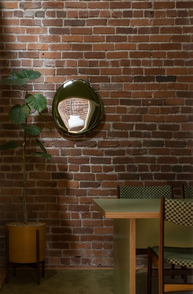 A moody dining area with an exposed red brick wall, dark green flooring, a small green-topped dining table, and patterned upholstered dining chairs. A tall potted plant and rounded reflective wall mirror add organic and sculptural accents. The space has a warm, intimate, vintage-inspired feel with industrial architectural detailing.