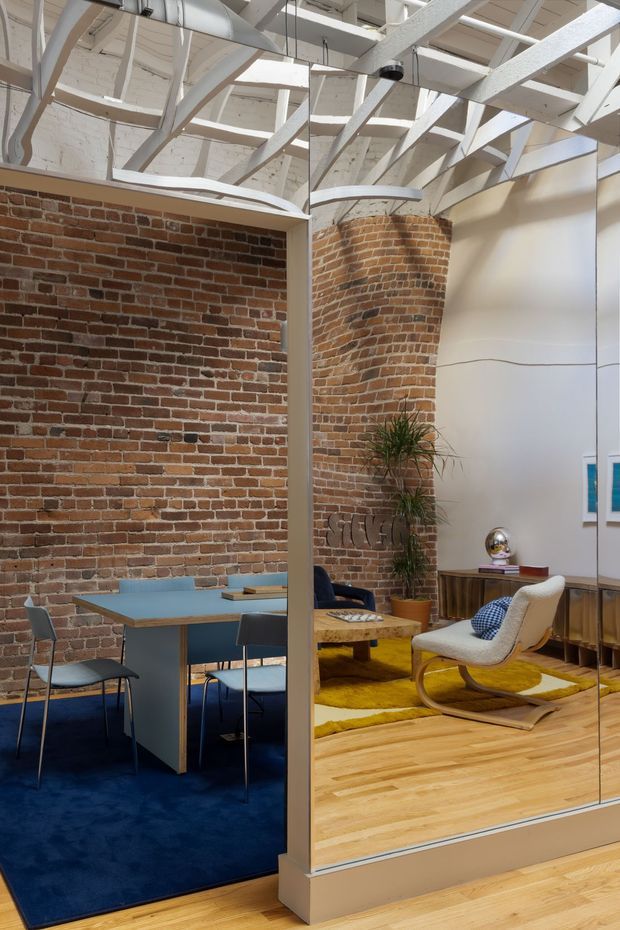 A loft-like interior with exposed red brick walls, a white-painted open timber and steel roof structure, and a glazed partition dividing a dining or meeting area from a lounge. The space features timber flooring, deep blue carpet, a blue dining table with chairs, a yellow rug, a curved lounge chair, indoor planting, and restrained mid-century furnishings. The visible structure and material palette suggest an adaptive industrial conversion with a warm, curated interior.