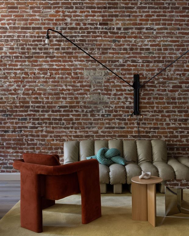 A moody, eclectic living room with an exposed red brick feature wall, a large tufted beige sectional sofa, and a rust-red sculptural armchair. A black articulated wall lamp spans the brickwork, while a small round timber side table and reflective metal table sit on a muted beige rug. The space combines industrial architectural fabric with distinctive contemporary furniture and playful patterned cushions.