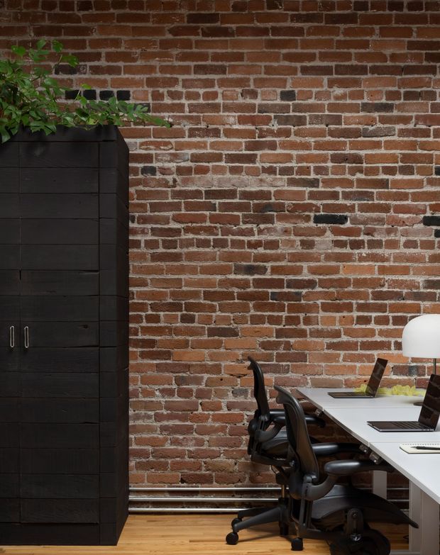 A compact industrial-style workspace with an exposed red brick wall, warm timber flooring, a long white desk, black ergonomic office chairs, laptops, and a dark built-in storage cabinet topped with a leafy plant. A white desk lamp adds a bright accent.