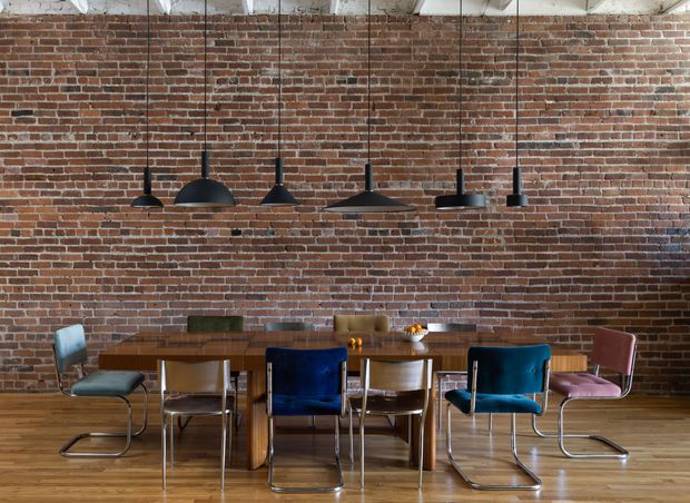 A loft-like dining area with a long wooden dining table and assorted upholstered cantilever chairs, set against an exposed red brick wall. Several black pendant lights hang at varied heights above the table, while the timber floor and exposed white ceiling structure reinforce the industrial character.