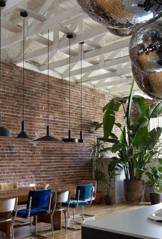 A dramatic dining area in an adaptive industrial-style interior, featuring an exposed red brick wall, white-painted timber roof trusses and a high ceiling with skylight-like glazing. A long timber dining table is paired with black pendant lights and colourful velvet dining chairs. Large tropical indoor plants and oversized mirrored disco-ball pendants add a playful, eclectic character. A grey kitchen island or benchtop is visible in the foreground.
