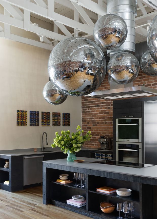 A dramatic loft-style kitchen with black fixed cabinetry, a large island with open shelving, integrated stainless-steel appliances, a dark benchtop, and a brick feature wall. White-painted exposed timber roof trusses and exposed ductwork create an industrial architectural character, while multiple oversized mirrored disco-ball pendant lights add a playful reflective feature. Timber flooring, colourful wall art, and floral styling complete the space.