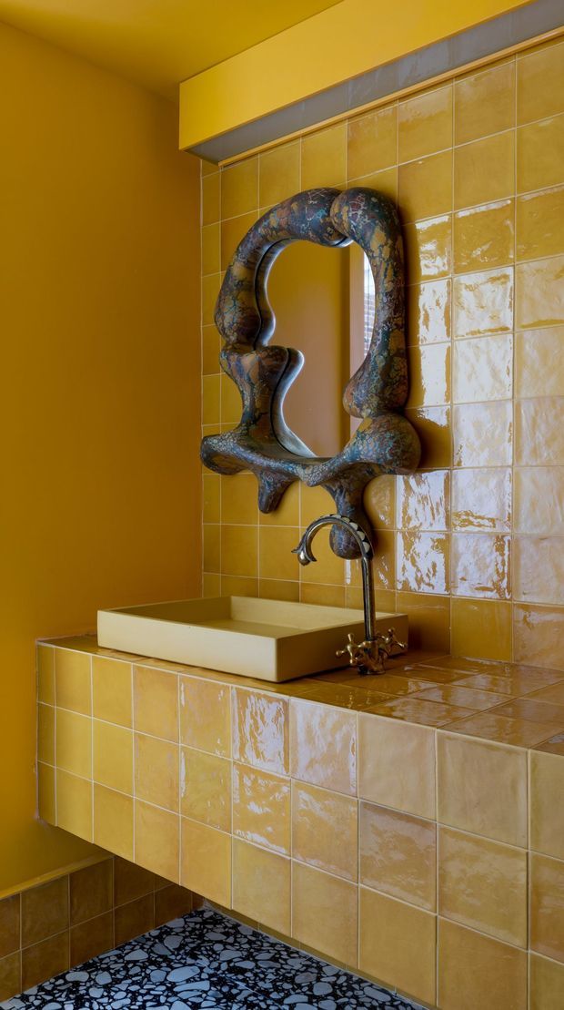 A compact, richly coloured bathroom vanity area with mustard-yellow painted walls and glossy yellow ceramic tile covering the backsplash, benchtop, and floating vanity front. A cream rectangular vessel basin sits beneath an ornate sculptural dark-framed mirror, paired with an aged bronze or dark metal tap. The floor features a bold black terrazzo-like pattern with light aggregate. The image has a playful, dramatic, highly curated interior aesthetic.