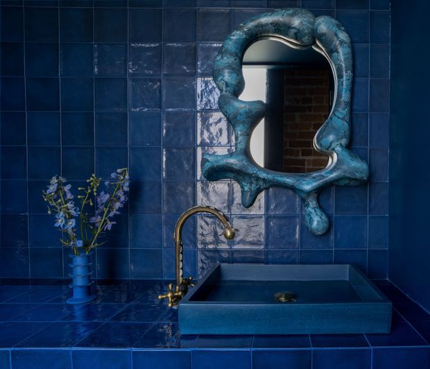 A compact, moody bathroom vanity clad entirely in glossy deep-blue square tiles. A sculptural irregular blue-framed mirror hangs above a rectangular integrated basin, paired with an aged brass wall-mounted faucet. A small blue vessel with delicate branches sits on the tiled counter, while a brick-lined space is reflected in the mirror.