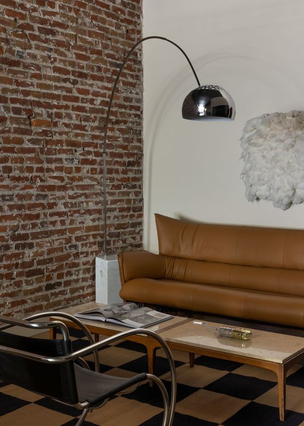 A stylish living room with an exposed red brick wall, warm tan leather sofa, black leather-and-chrome lounge chairs, and a low rectangular coffee table. A dramatic arched floor lamp with a reflective dome shade hangs over the seating area, while a white sculptural wall piece adds texture. The room has a bold black-and-tan checkerboard rug and a refined, loft-like atmosphere.