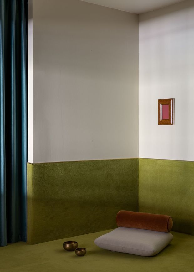 A moody, stylised interior corner with olive-green textured wall panelling continuing onto the floor, warm off-white upper walls, and deep teal curtains at the left. A low sculptural lounge cushion with a rust-brown cylindrical bolster sits beside two small bronze-toned bowls. A small framed artwork adds a pink accent on the wall.
