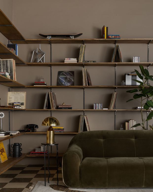 A moody, carefully styled living room focused on a full wall of open timber shelving supported by dark metal framing. The shelves display books, vinyl records, decorative objects, and a skateboard, with a large deep olive-green upholstered sofa in the foreground. A brass table lamp, black side tables, a checkerboard floor, and a leafy indoor plant contribute to the richly layered, eclectic interior.