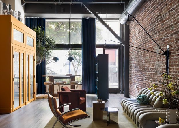 A stylish industrial loft living room with exposed red brick walls, a high dark ceiling with visible ductwork and beams, large black-framed windows, and dark curtains. The space includes a sculptural upholstered sofa, leather lounge chairs, a small coffee table arrangement, indoor plants, and a tall timber-framed glazed storage cabinet. A compact dining or work area is positioned near the windows.