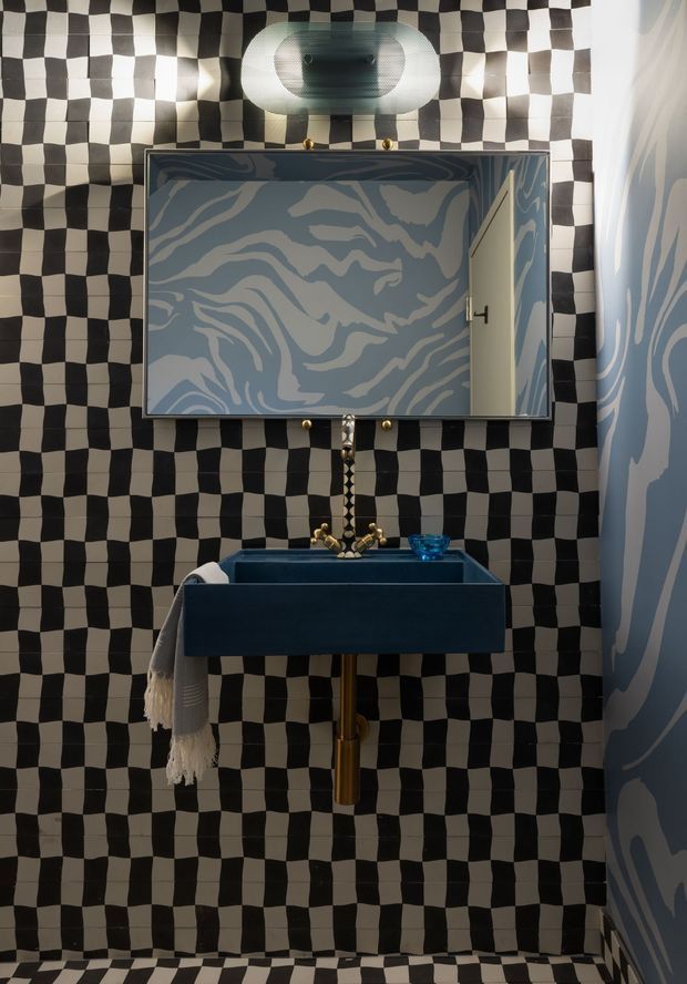A compact, highly stylised bathroom or powder room featuring a wall-mounted deep blue basin with brass plumbing and tapware, a large mirrored cabinet, and a sculptural wall light. The walls and floor are covered in a bold black-and-white checkerboard tile pattern, while the mirror cabinet and adjacent wall feature blue-and-pale abstract organic patterned finishes. A towel hangs from the basin, and the space has dramatic, playful decorative detailing.