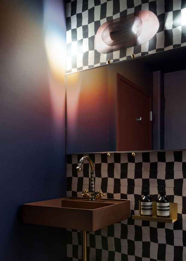 A compact, dramatically lit bathroom vanity featuring a dark navy wall, a copper-toned rectangular basin with brass plumbing and tapware, and a mirrored cabinet. The backsplash and upper wall are covered in bold black-and-white checkerboard tiles, complemented by a sculptural wall light and a small brass shelf holding dispensers. The interior has a moody, theatrical, highly graphic character.