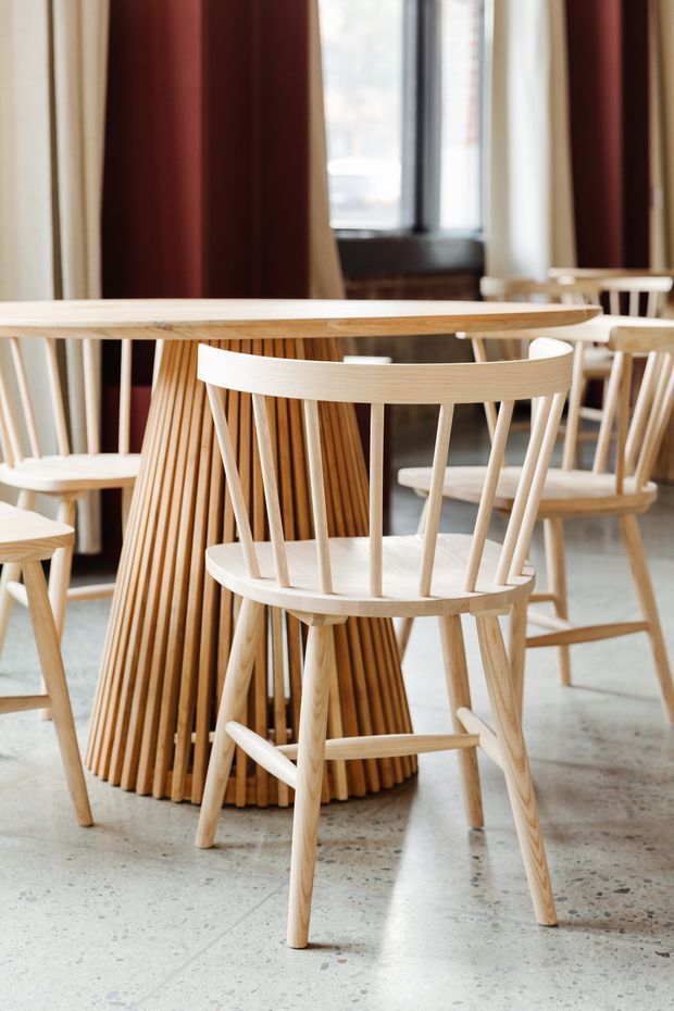 A warmly styled dining area featuring a round light timber dining table with a sculptural slatted pedestal base, surrounded by matching pale timber spindle-back chairs. The space has a polished concrete or terrazzo-like floor, tall windows with dark frames, and layered cream and deep burgundy curtains. The image focuses on natural timber furniture and a refined, contemporary interior setting.