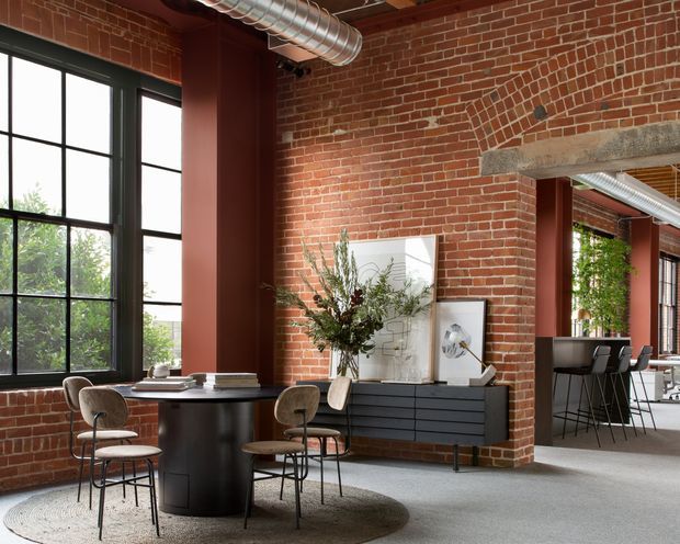 A loft-style dining and living space with exposed red brick walls, tall black-framed industrial windows, exposed galvanized ductwork, and a concrete lintel over a wide opening. A round dark dining table with upholstered chairs sits beside a low black sideboard, framed artwork, and a large vase of branches. A bar or kitchen area with stools is visible through the opening. The room has a warm, refined industrial character with abundant natural light and a high ceiling.