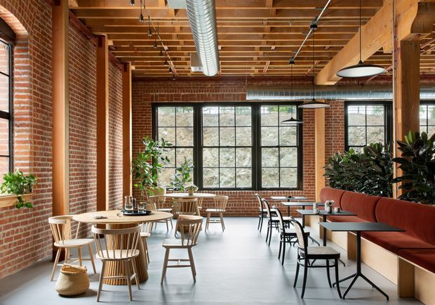 A spacious industrial-style café or hospitality interior with exposed red brick walls, timber post-and-beam structure, open timber ceiling joists, visible metal ductwork, black-framed grid windows, and polished concrete flooring. The room is furnished with round timber tables, spindle-back chairs, black metal café tables, and a long rust-coloured upholstered banquette. Pendant lighting and indoor plants add warmth to the otherwise robust material palette.