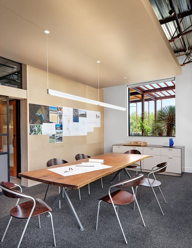 Bright contemporary design studio or meeting room with a large timber worktable surrounded by mid-century style chairs. The room has dark carpet flooring, pale timber wall panelling, white built-in drawer cabinetry, a suspended linear pendant light, and architectural drawings pinned to the wall. Black-framed glazing opens the space to a covered outdoor area with planting visible beyond.