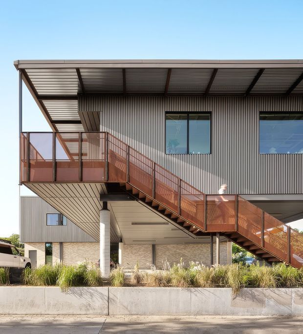 Exterior architectural photograph of a contemporary elevated building with corrugated metal cladding, a broad low-pitched roof canopy, large windows, exposed structural columns, and a prominent external stair with rust-coloured perforated metal balustrades. A landscaped concrete planter occupies the foreground.