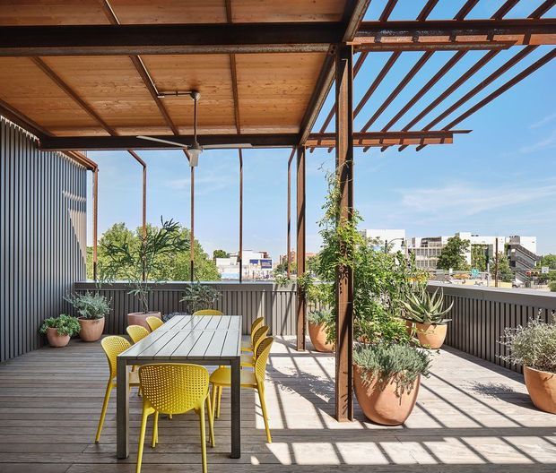A bright rooftop or elevated outdoor dining terrace with a covered timber pergola and exposed steel supports. A long outdoor dining table is surrounded by yellow chairs, while corrugated metal privacy screens, timber decking, climbing plants, and large potted plants frame views toward an urban setting. Strong geometric shadows create a warm, contemporary atmosphere.