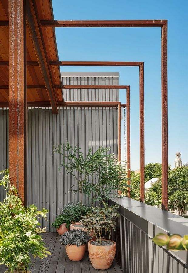 A sunny outdoor balcony or terrace with dark timber decking, corrugated metal privacy cladding, and a series of rusted steel rectangular frames forming a pergola-like architectural structure. Potted plants line the edge, with a leafy suburban outlook and a distant tower visible beyond the railing.