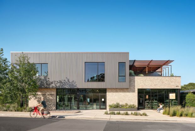 Street-facing contemporary commercial building with a two-storey composition. The upper level is clad in vertically ribbed grey metal, while the lower level combines expansive dark-framed glazing with pale masonry cladding. A timber pergola and planted terrace sit above the right-hand frontage. Landscaping, footpath, and street activity frame the building exterior.