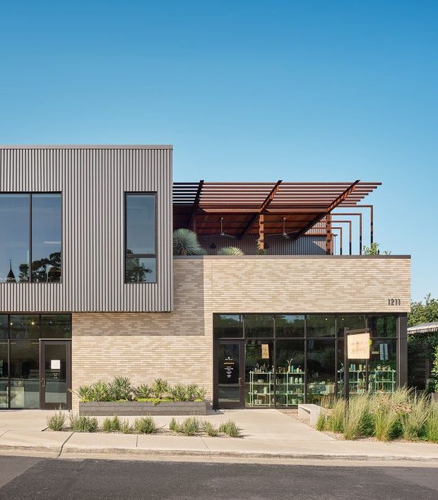 Street-facing contemporary commercial building with a two-storey facade combining vertically ribbed metal cladding, light brickwork and extensive dark-framed glazing. A timber pergola shades an elevated terrace, while landscaped planting borders the pedestrian frontage.