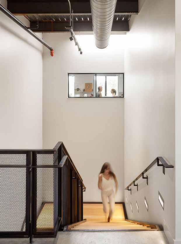 A tall, narrow interior stairwell with a straight timber staircase descending toward the camera. The space has white plaster walls, black metal mesh balustrades and handrails, exposed dark ceiling structure, visible ductwork and track lighting, and a small internal window looking into an adjacent room. The design has a clean industrial character with strong vertical volume and bright natural light.