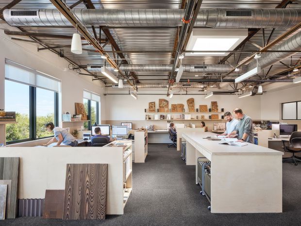 A bright open-plan commercial design studio or office with exposed metal ductwork, structural beams, conduit, and corrugated ceiling panels. Large black-framed windows line the left wall, while white workstations, desks, computers, shelving, material samples, and a central island support collaborative architectural or design work. The space has an industrial architectural character softened by clean contemporary furnishings and abundant daylight.