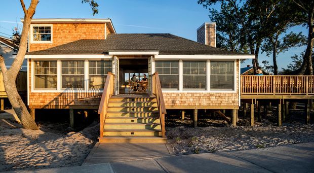 Exterior front view of a raised coastal-style timber home with cedar shingle cladding, a dark hipped roof, broad screened or glazed porch windows, and a central staircase leading to the elevated entry. A timber deck extends to the right, with mature trees and sandy ground surrounding the structure.