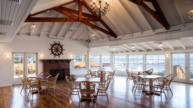 Bright coastal dining hall with large windows overlooking a marina, featuring a white-painted vaulted timber ceiling with exposed dark wood trusses, polished timber flooring, a central brick fireplace, nautical ship-wheel wall decor, chandeliers, and numerous round dining tables with wooden spindle-back chairs.