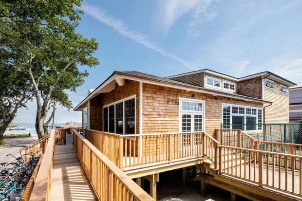 Exterior view of a raised coastal-style house with extensive timber decking, ramps and balustrades. The facade is clad in small wood shingles, with white-trimmed multi-pane windows, a low-pitched roof and dormer windows. A waterfront or marina-like setting is visible at the side, with bicycles and neighbouring buildings in the background.