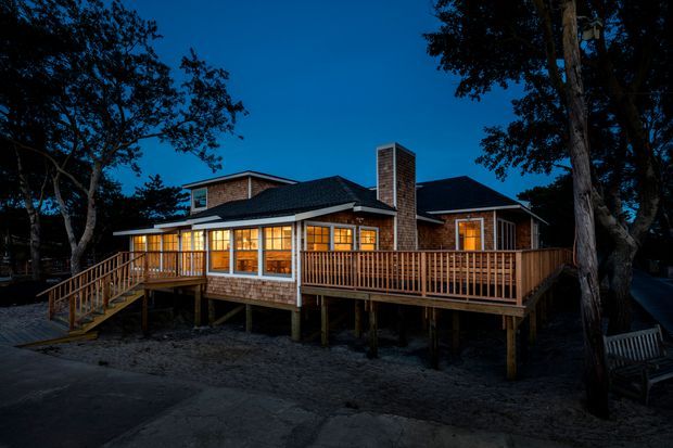 A warmly illuminated single-storey brick-clad house photographed at dusk, elevated above the ground on timber posts. The home has multiple dark pitched roof forms, a prominent brick chimney, extensive windows, and a large timber deck with balustrades and stairs, surrounded by mature trees.