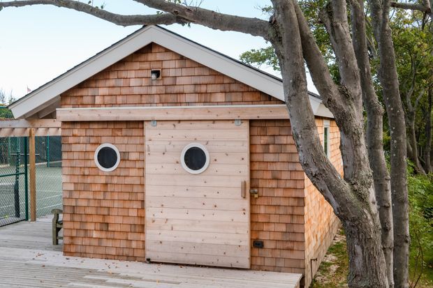 Exterior view of a small, single-storey timber-clad outbuilding with a simple gable roof. The facade is covered in warm cedar shingles, with pale timber trim and a distinctive double circular-window door. It sits beside a timber deck and fenced outdoor area, partially screened by mature trees.
