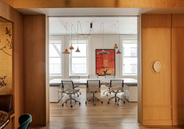 View through a timber-lined opening into a bright, compact office or studio. The workspace has a pale painted ceiling and walls, three desks with wheeled task chairs, white drawer units, large multi-pane windows, and a central red artwork. Multiple suspended pendant lights hang from exposed cords. Warm wood wall panelling and built-in cabinetry frame the office, with a decorative niche visible at left.