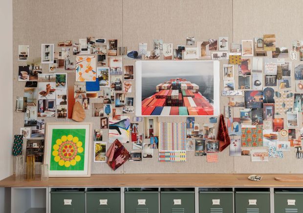 A styled office or creative workspace wall covered with a dense collage of photographs, artwork, postcards, magazine cuttings, and inspiration images. A large framed photograph of colourful shipping containers is centrally mounted, with open shelving and display niches integrated into the collage. Below, a long timber ledge sits above rows of dark green storage drawers with label holders. The overall composition is eclectic, highly curated, and colourful against a neutral textured wall.
