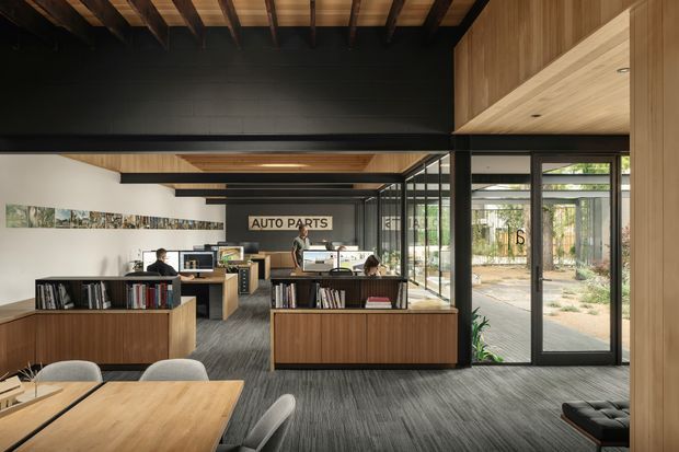 A contemporary open-plan commercial office or showroom with timber-clad built-in counters and walls, exposed dark timber ceiling beams, black structural elements, glass partitions, and abundant glazing opening toward a landscaped courtyard. Workstations, shelving, desks, and a meeting table are visible, with an “AUTO PARTS” sign at the rear.