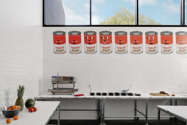 Bright commercial-style kitchen with white subway-tile walls, stainless-steel worktables and commercial espresso equipment. A row of tomato soup cans forms a bold decorative graphic above the preparation bench. High black-framed windows bring in daylight and show trees outside, while fruit and kitchen equipment occupy the foreground and counters.