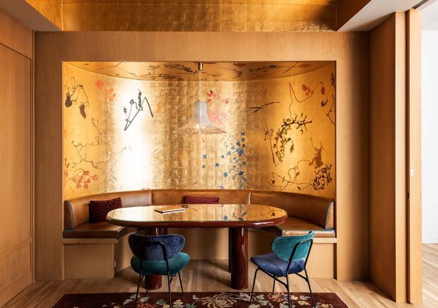 A richly detailed built-in dining nook with a curved banquette, oval dining table, and two sculptural chairs. Warm timber wall panelling and cabinetry frame a large metallic gold-toned decorative wall panel featuring botanical and abstract linework. The space has integrated lighting, timber flooring, and a refined mid-century-inspired atmosphere.