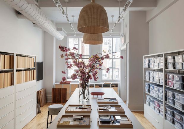 Bright, contemporary design showroom or studio with a long central worktable displaying material samples, shelving of organised sample books and products, large windows, exposed white services, track lighting, and woven pendant lights. A floral branch arrangement adds a soft decorative focal point.