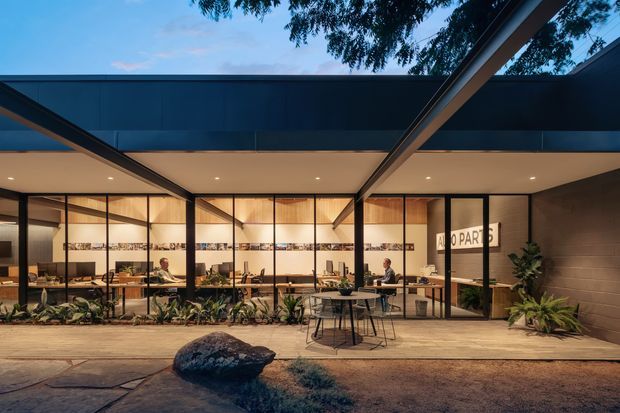 Twilight exterior view of a contemporary commercial pavilion with broad dark metal roof forms, warm recessed lighting, and extensive floor-to-ceiling glazing. The glazed frontage reveals an open office or hospitality-style interior with desks, tables, seating, pendant lights, and plants. A covered paved forecourt with integrated landscaping and a large feature rock occupies the foreground.