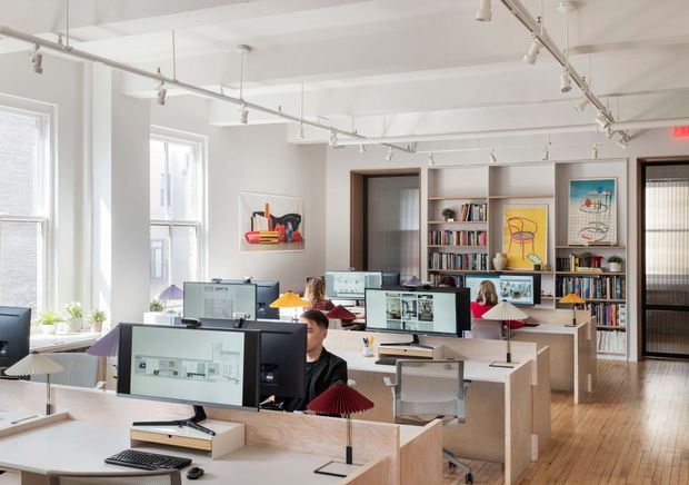 Bright open-plan creative studio or office with multiple individual workstations, computer monitors, desk lamps, shelving filled with books and objects, framed artwork, and timber flooring. White walls and exposed ceiling services are paired with pale timber desks and abundant daylight from large windows. A glazed or screened doorway and corridor appear at the rear.