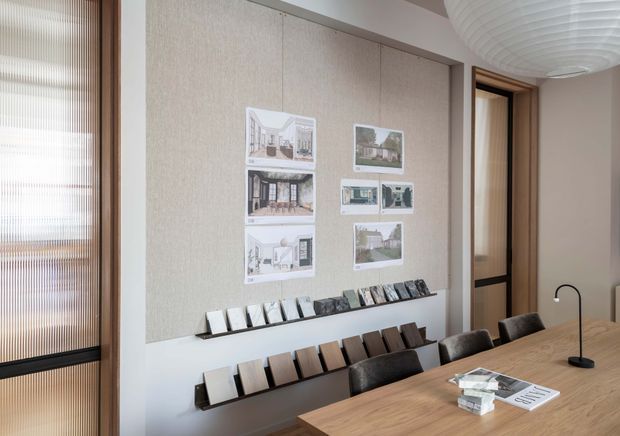 A refined interior design presentation and dining area. A large textured neutral wall panel displays architectural concept images above rows of material samples, while a long timber dining table with dark upholstered chairs occupies the foreground. Warm timber-framed glazing and a sculptural white pendant light add natural, understated detail.