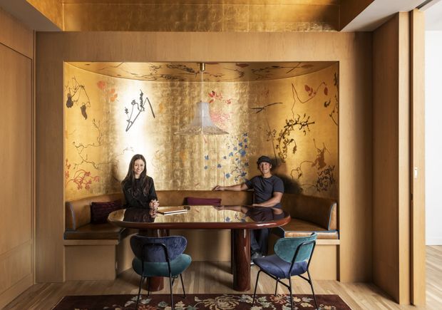 A richly detailed dining or lounge nook framed by warm timber joinery, featuring a built-in brown leather banquette, round polished table, and two upholstered chairs. The alcove is wrapped in a dramatic gold-toned decorative mural or metallic artwork with botanical and abstract motifs. Warm integrated lighting, timber flooring, and an adjacent bright opening create a refined, intimate atmosphere.