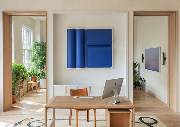 A bright, refined home office viewed frontally, featuring a light timber desk with an all-in-one desktop computer, a blue sculptural wall artwork, pale walls, timber flooring, and built-in low cabinetry. Open timber-framed doorways reveal a plant-filled adjoining room on the left and a hallway or gallery area on the right. A patterned rug adds a subtle decorative element.