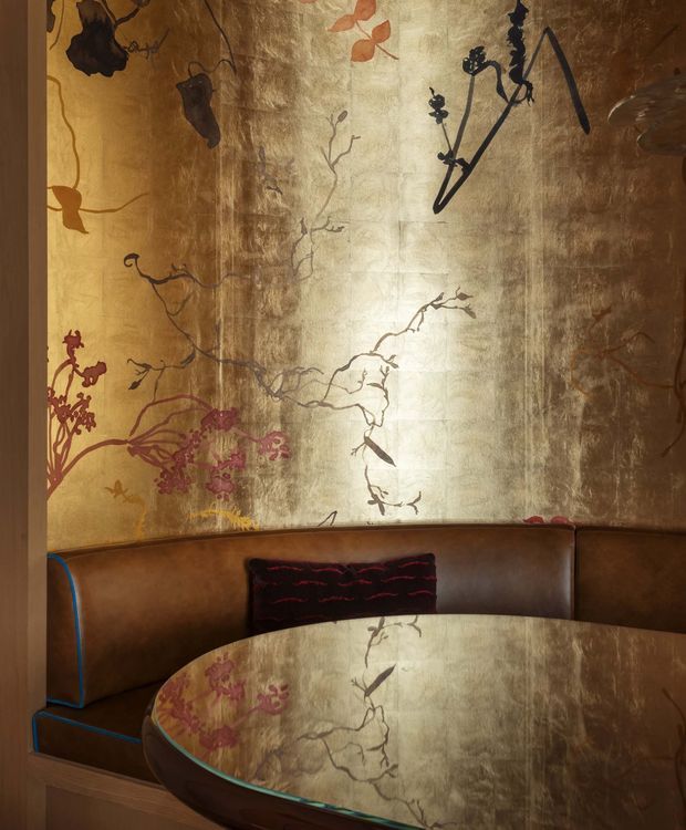 A richly detailed intimate dining nook with a curved brown leather banquette, round reflective glass-topped table, and an ornate metallic wall treatment featuring botanical and branch-like artwork. Blue piping accents the banquette, while warm bronze, brown, and gold tones create a dramatic, luxurious atmosphere.