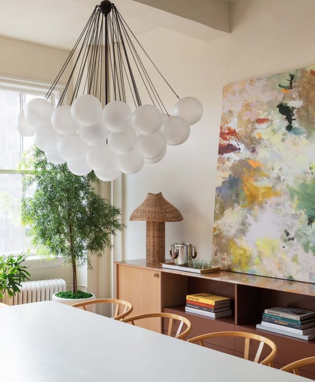 Bright, refined dining area with a large white dining table and timber-framed chairs. A sculptural pendant light made of clustered white globes hangs from multiple dark cords above the table. The room features a warm timber sideboard with open shelving, books and decorative objects, a large colourful abstract artwork, indoor plants, a radiator, and generous window light.