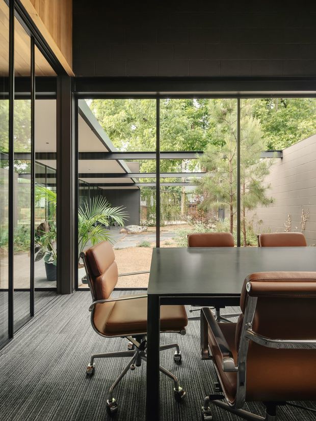 A refined contemporary office or meeting room with a large dark rectangular conference table and brown leather office chairs. Full-height black-framed glazing opens the room visually to a landscaped courtyard, while exposed black masonry walls, timber ceiling lining, and a covered outdoor structure create a strong indoor–outdoor connection. The room has carpet flooring and abundant natural light.