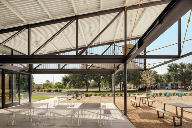 A spacious open-air pavilion or covered terrace with exposed dark steel framing, a high white ribbed metal roof, extensive glazing, and outdoor café-style tables and benches. The space opens onto a landscaped courtyard with mature trees and pathways, creating a bright, contemporary communal setting.