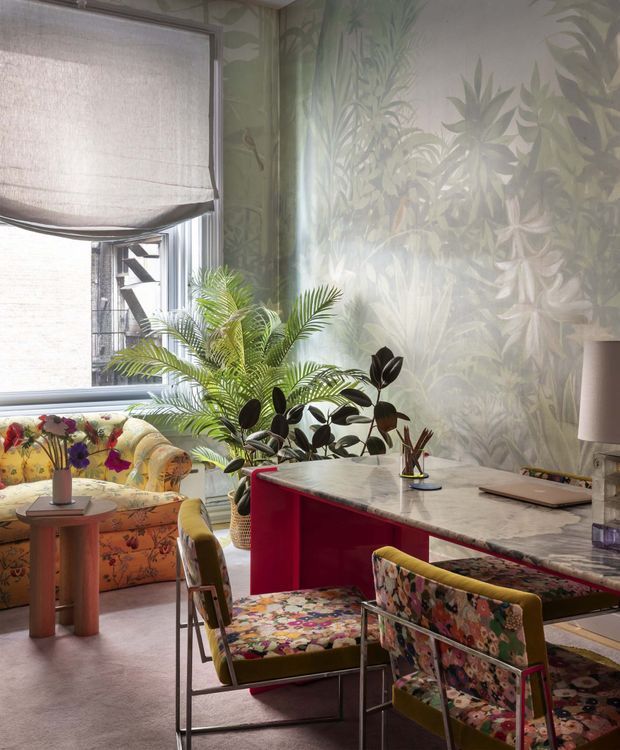 A richly patterned, plant-filled interior combining a compact dining or work area with a vivid floral sofa by a large window. A marble-topped curved peninsula or desk with a deep red base dominates the foreground, accompanied by colourful floral-upholstered metal-framed chairs. Botanical wallpaper, indoor palms, a roller blind, table lamp, and layered floral fabrics create an eclectic, maximalist atmosphere with strong natural light.