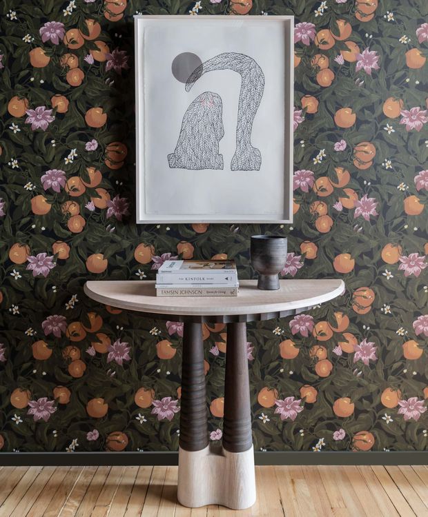 A styled interior vignette featuring a narrow curved console table with sculptural two-tone timber legs, placed against richly patterned dark floral wallpaper. Books and a dark metallic vessel sit on the console, beneath a framed monochrome artwork. Timber floorboards are visible at the base. The scene has an eclectic, decorative, gallery-like character.