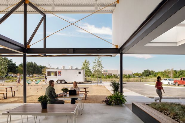 Open-air contemporary pavilion or commercial courtyard with a covered terrace, exposed dark steel framing, diagonal tension cables, corrugated roof panels, and large open glazed or unglazed bays. Café-style tables and seating occupy the concrete floor, with built-in raised planters and ornamental grasses. The space looks out toward a landscaped suburban or urban setting with a food trailer and parking beyond.