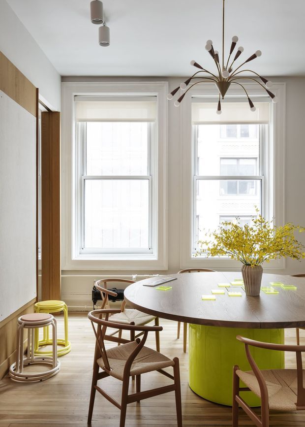 Bright, stylish dining area in a renovated interior, featuring a large oval wood dining table with a vivid lime-green cylindrical base, woven-seat timber dining chairs, and a sculptural mid-century-inspired pendant light. Two tall sash windows with white trim bring in abundant daylight. Pale timber flooring, white walls, and a slim timber-and-white built-in element create a clean, contemporary setting, with a vase of yellow foliage as a tabletop centerpiece.
